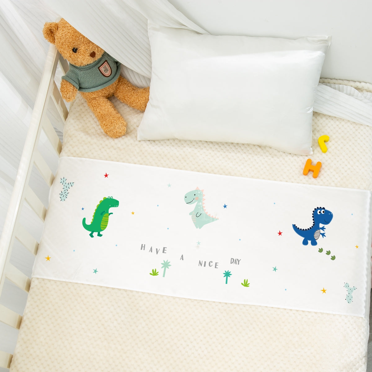A white crib bumper with dinosaur print and 'HAVE A NICE DAY' text in a baby crib interior with stuffed bear and alphabet blocks.