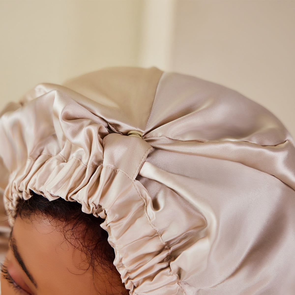 A beige mulberry silk sleep cap worn by a young woman, showcasing its smooth texture and gathered fit against a plain studio backdrop.