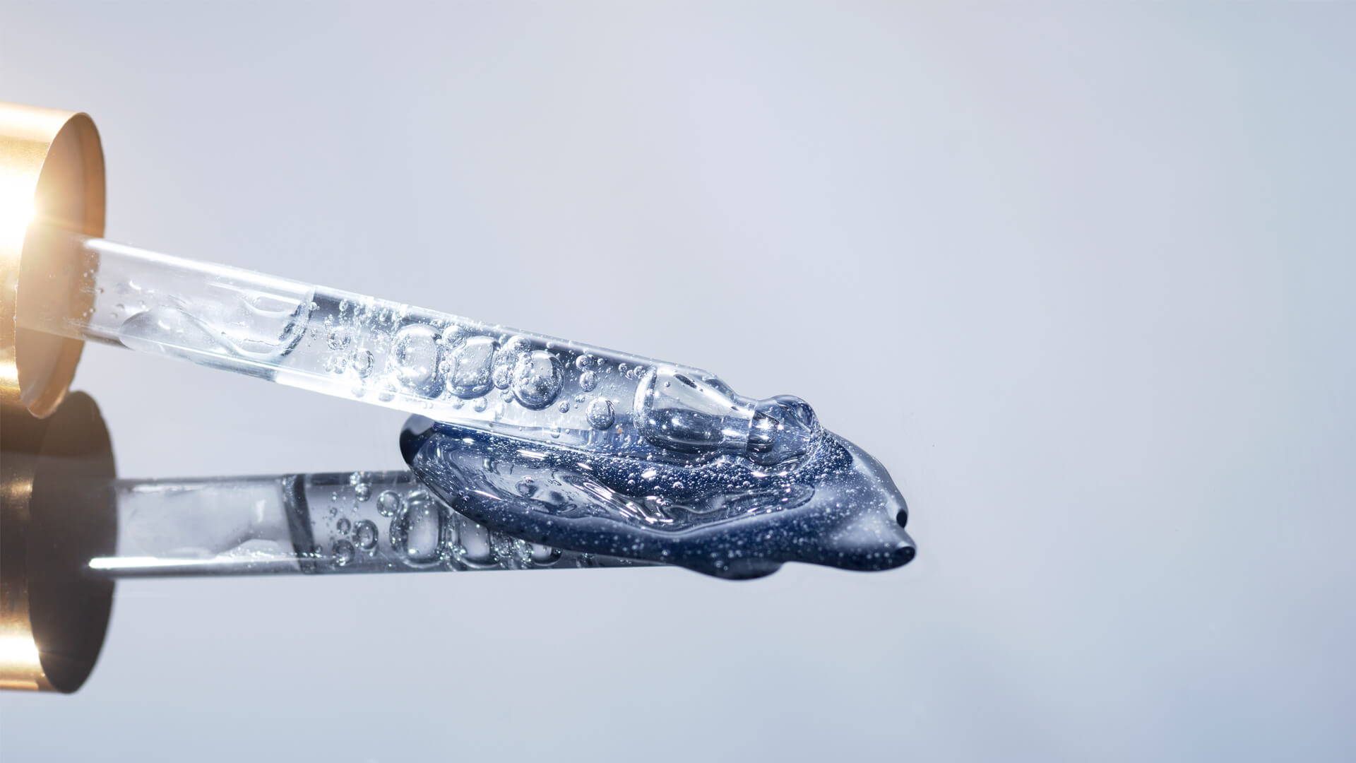 A clear gel-like substance with a blue tint being dispensed from a gold-tipped tube on a light gray studio backdrop.
