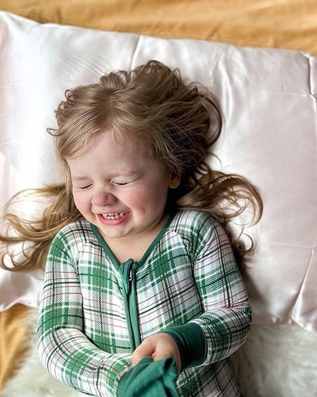 A young child in a green and white plaid pajama top lies on a bed with a white pillow and yellow bedding, laughing and playing.
