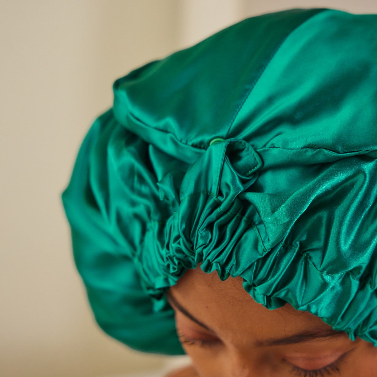 A woman wearing an emerald green mulberry silk bonnet over her hair, with a plain studio backdrop.