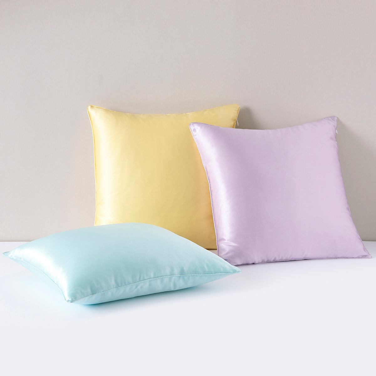 Three mulberry silk pillowcases in yellow, lavender, and light blue arranged in a flat lay on a plain studio backdrop.