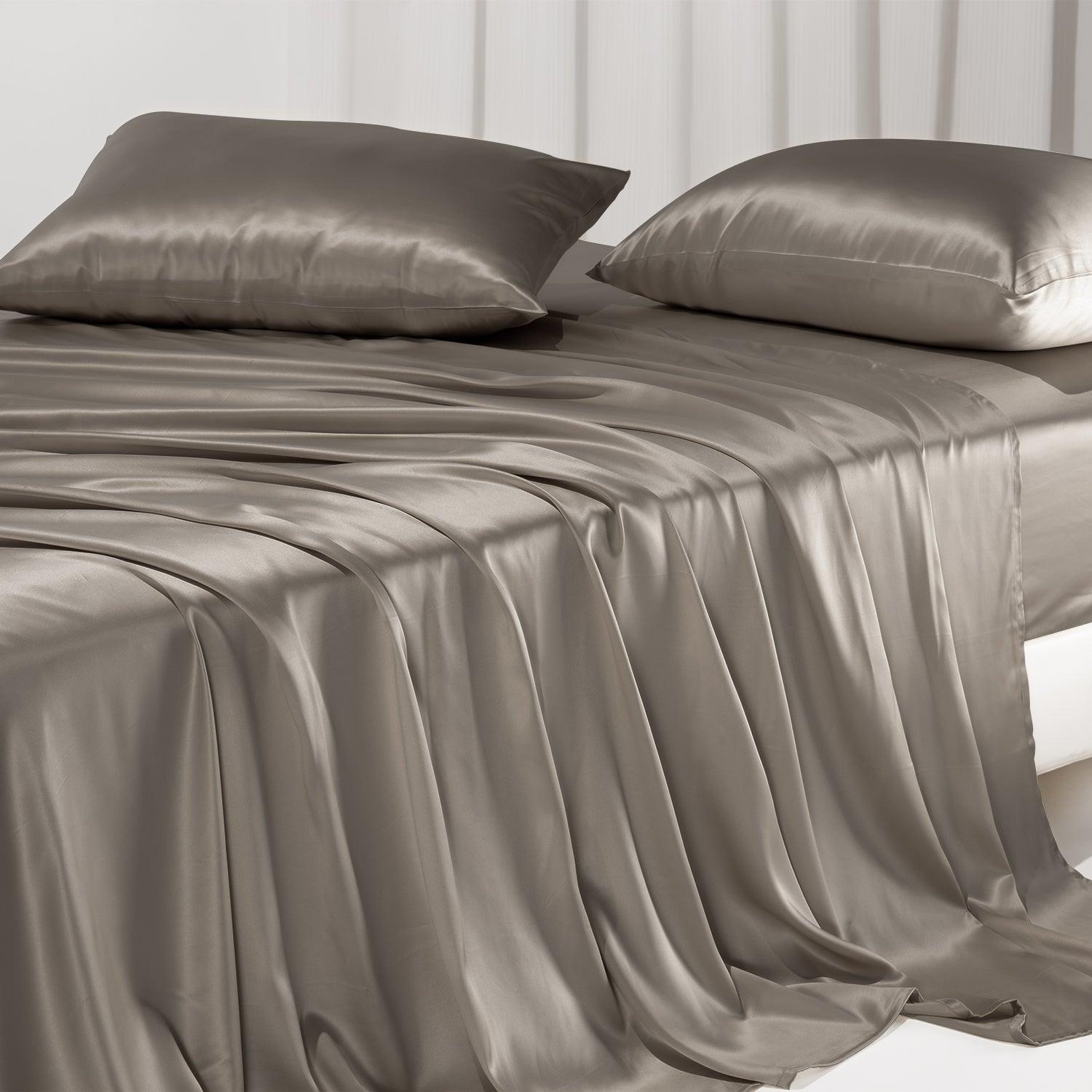 A taupe mulberry silk sheet set draped on an unmade bed with a white wall and vertical paneling, emphasizing texture and drape.