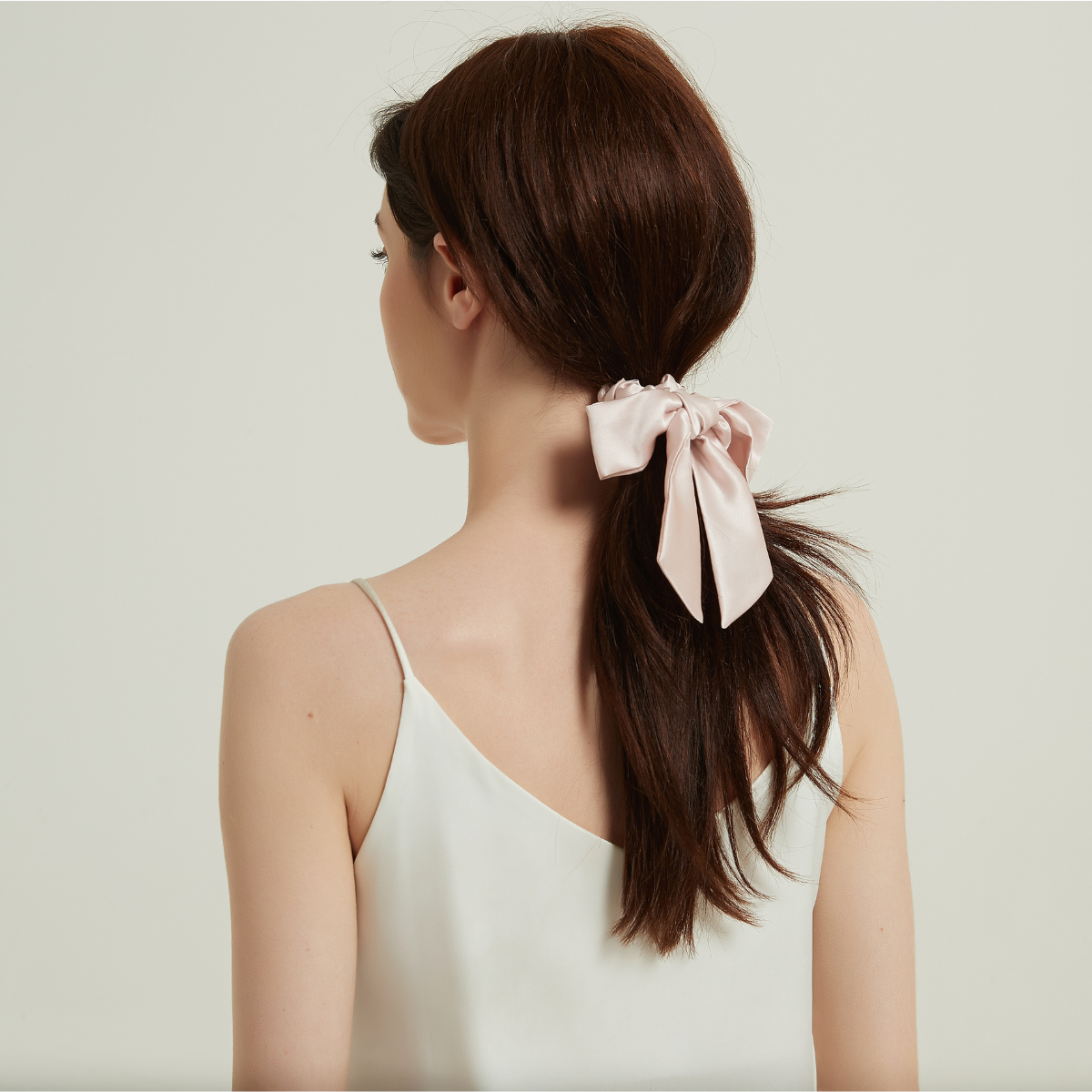 A pink mulberry silk hair bow tied in a ponytail on a woman with dark brown hair, against a plain studio backdrop.