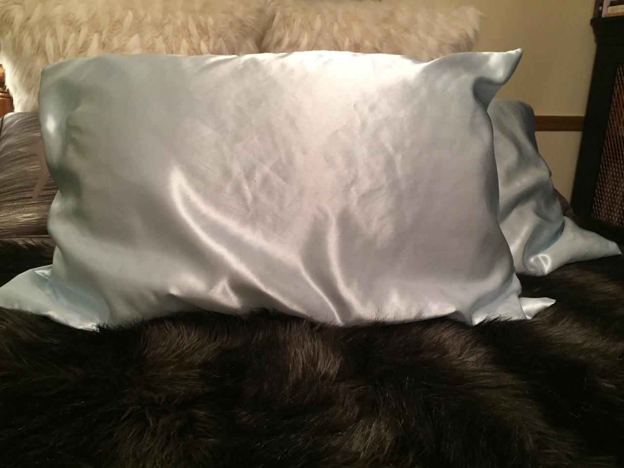 A light blue mulberry silk pillowcase on a bed with a dark fur blanket and fluffy pillows, slightly wrinkled for a casual look.