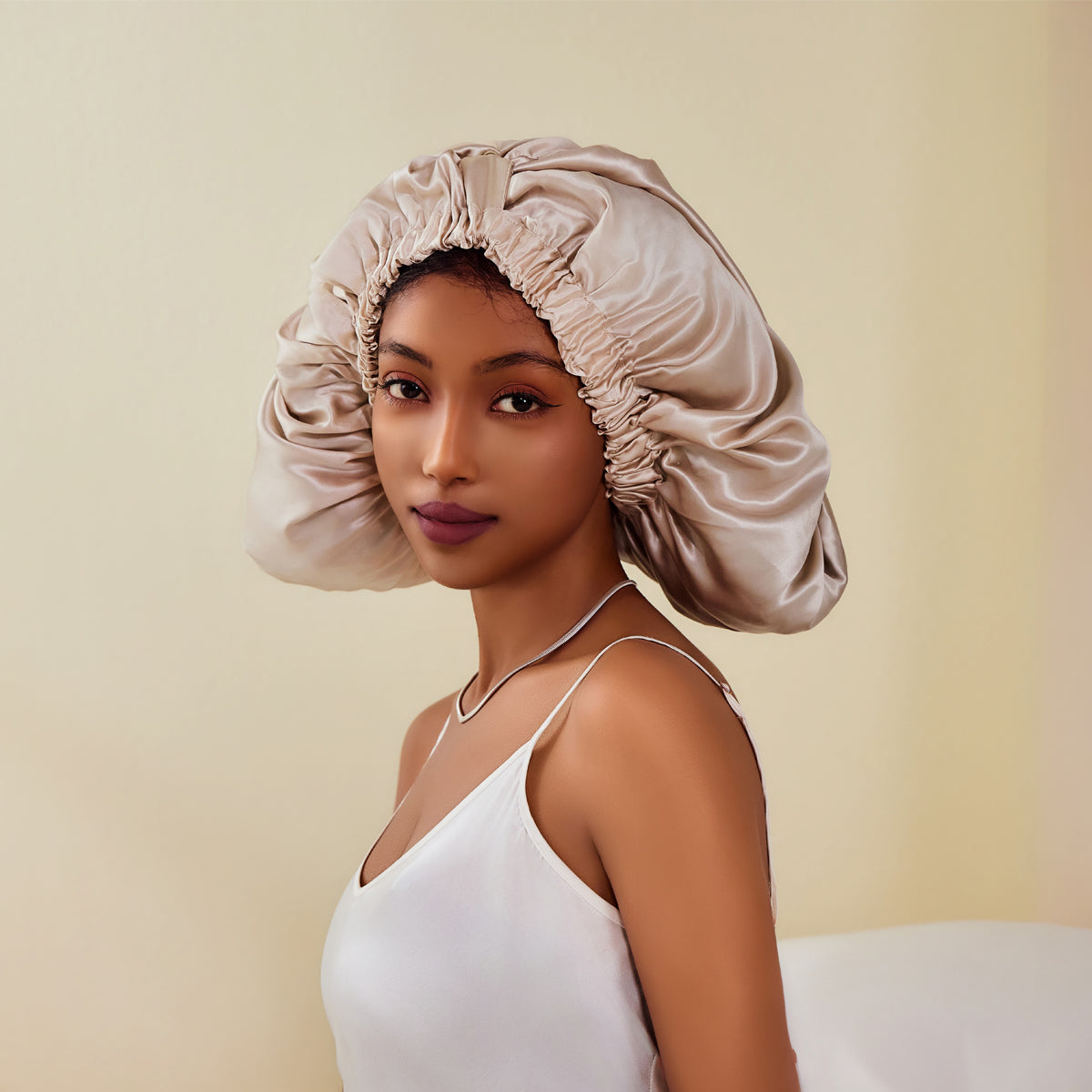 A beige mulberry silk sleep cap worn by a young woman with medium-dark skin tone against a plain studio backdrop. — silk sleep cap / bonnet