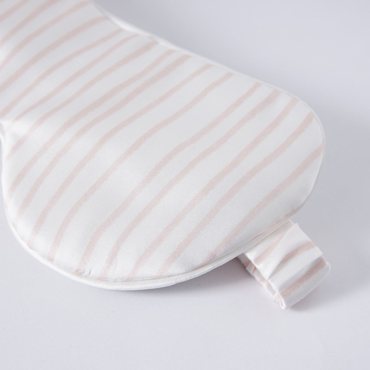 A white sleep eye mask with light pink stripes on a cream studio backdrop, folded strap visible on the side.