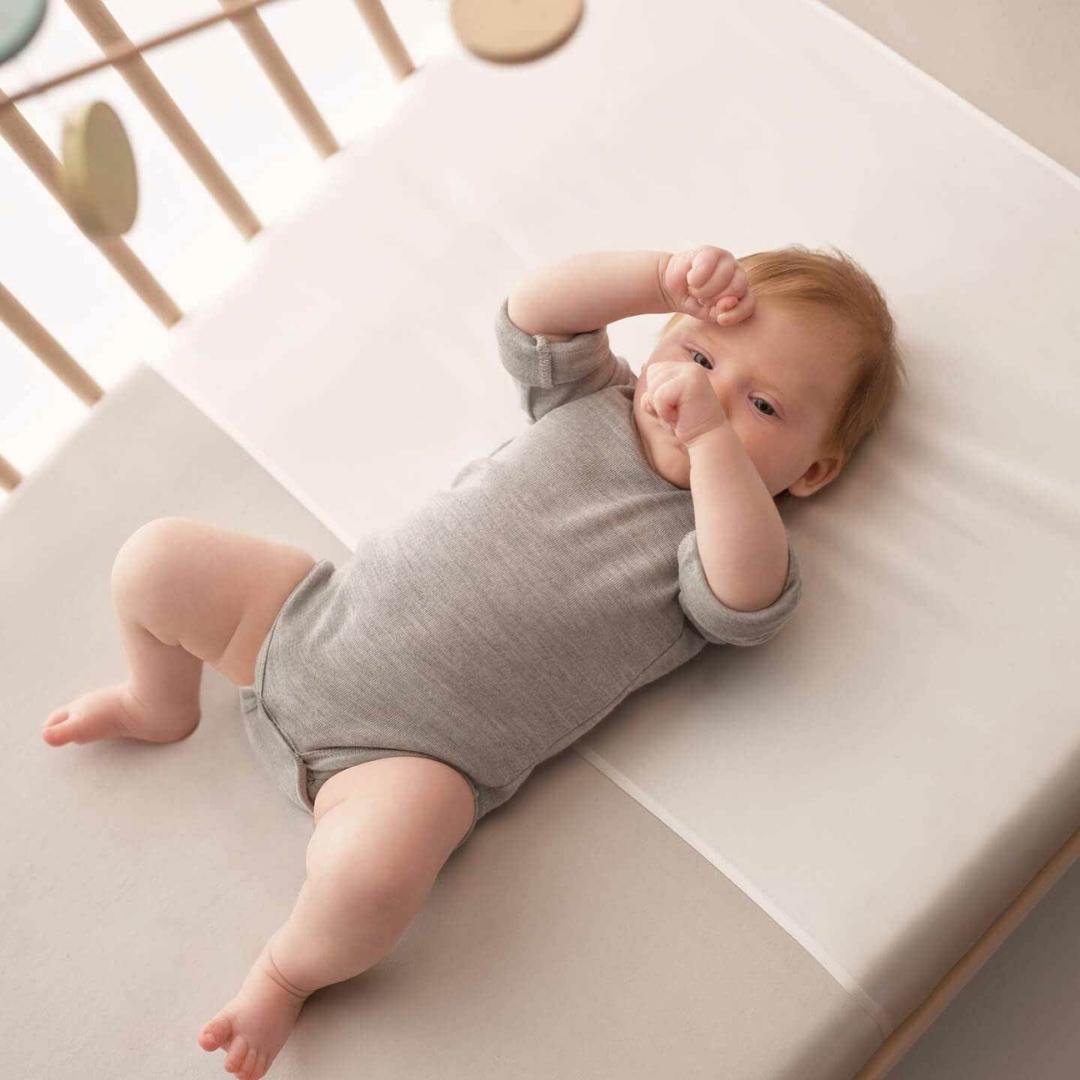 A baby wearing a gray long-sleeve onesie lies on its back in a crib with neutral bedding and wooden rails. — 5 Best Natural Sleep Aids: Whic